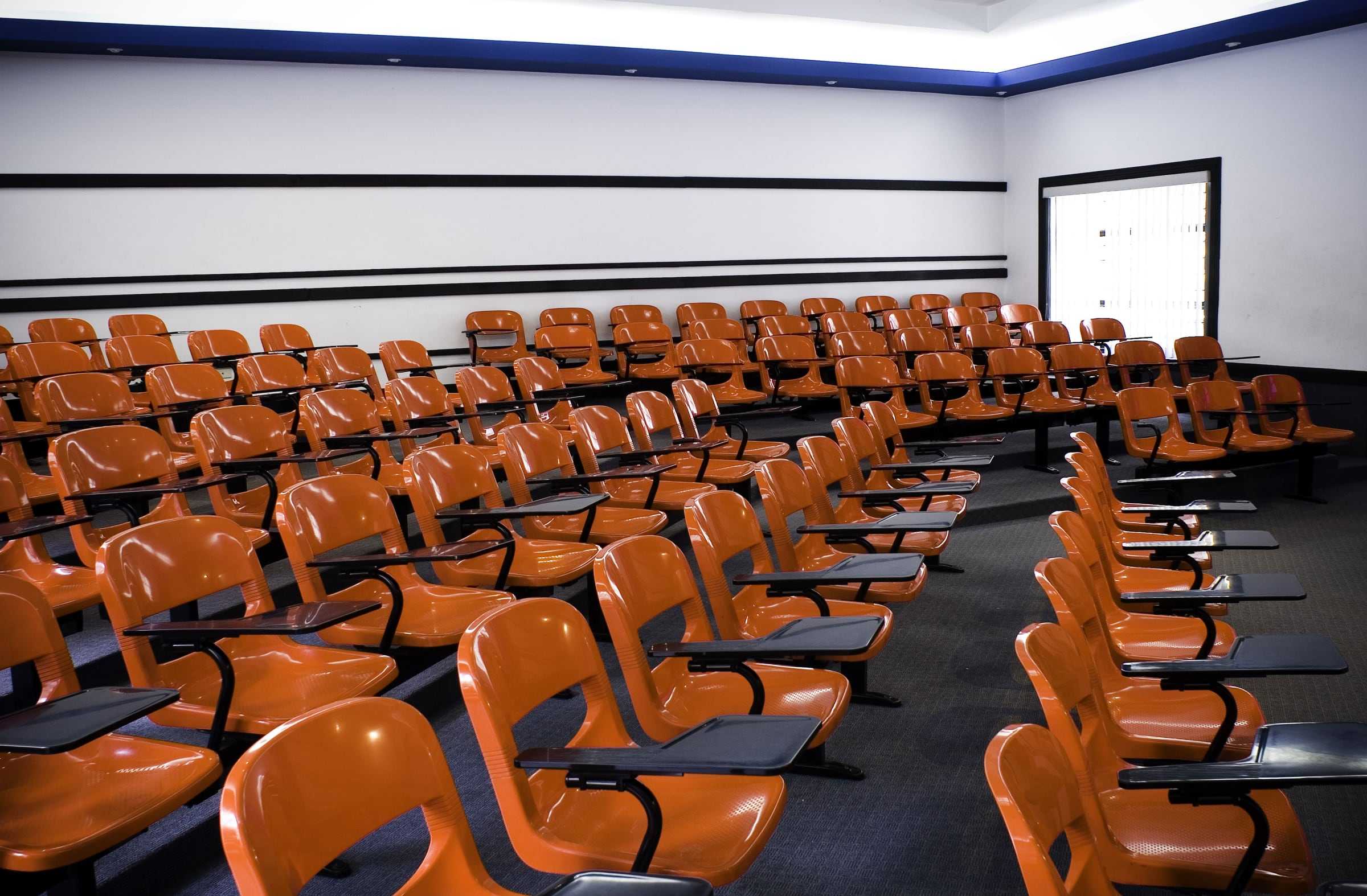 Auditorio de la Facultad de Arquitectura de la UCR con butacas anaranjadas con paleta en gradas, bajo una franja de luz azul